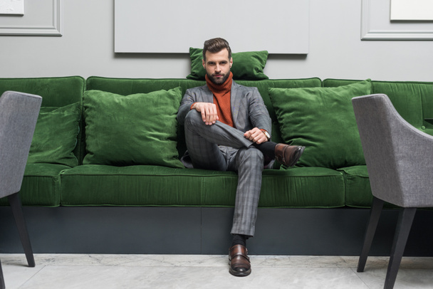 man in formal wear with legs crossed sitting on green sofa and looking at camera - Photo, Image