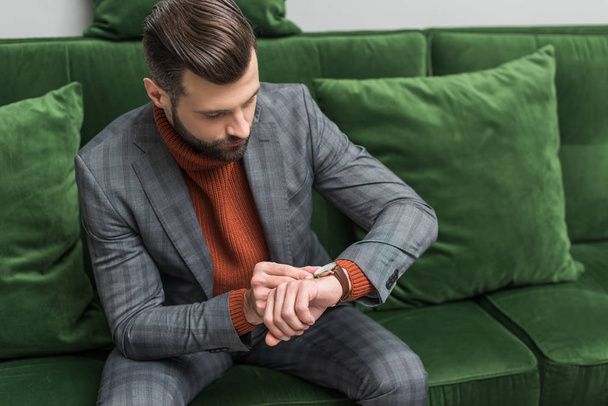 man in formal wear sitting on green sofa and adjusting watch on hand - Фото, зображення
