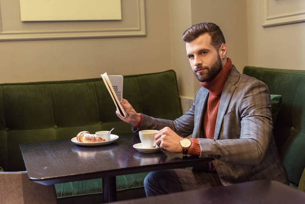 stylish man in formal wear sitting at table during lunch and reading newspaper in restaurant - 写真・画像