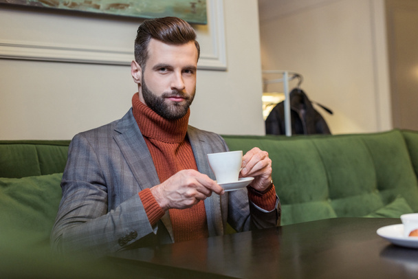 handsome man in formal wear looking at camera, sitting at table and drinking coffee in restaurant - Фото, зображення