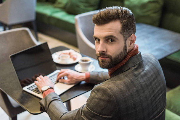handsome businessman sitting at table, looking at camera and typing on laptop with blank screen in restaurant - Foto, Imagem