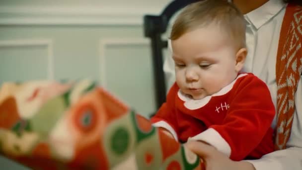 Amazing looked of A young girl gives a child a gift to a child who sits in her moms lap near a decorated Christmas tree and gift. Close UP - Materiał filmowy, wideo