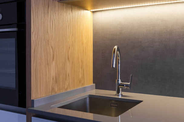 Un lavello sottopiano e un miscelatore montati su un piano in pietra ceramica in una moderna cucina soppalcata di colore grigio con dettagli in legno
.  - Foto, immagini