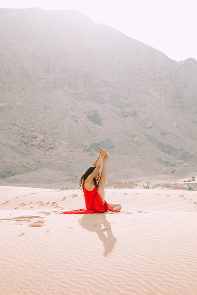 Fiatal gyönyörű nő, piros ruhás, jóga a sivatagban, a hegyek napkeltekor  - Fotó, kép