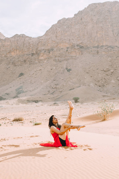 Fiatal gyönyörű nő, piros ruhás, jóga a sivatagban, a hegyek napkeltekor  - Fotó, kép