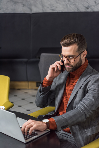 серьезный красивый бизнесмен в очках и формальной одежде сидит за столом, разговаривает на смартфоне и печатает на ноутбуке в ресторане
 - Фото, изображение