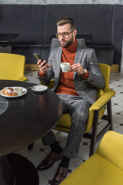 handsome man in formal wear using smartphone while drinking coffee in restaurant - Foto, afbeelding