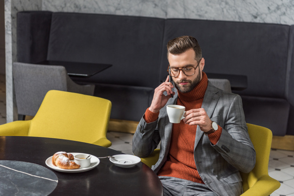 focused handsome man in formal wear talking on smartphone while drinking coffee in restaurant - Zdjęcie, obraz