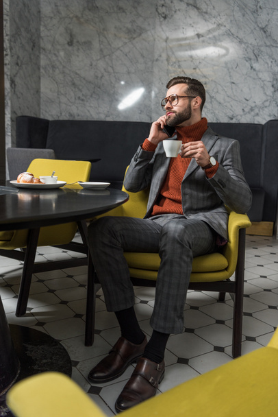 handsome man in formal wear talking on smartphone while drinking coffee in restaurant - Fotografie, Obrázek