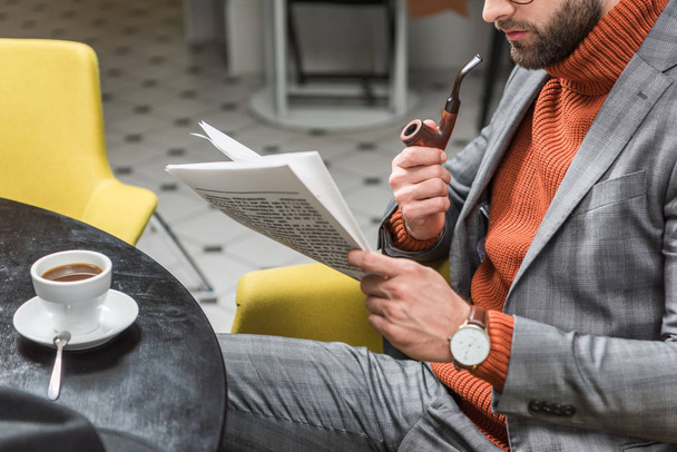 cropped view of stylish man in formal wear sitting at table, reading newspaper and smoking pipe in restaurant - Foto, immagini