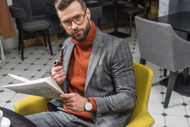 stylish man in formal wear looking at camera, reading newspaper and smoking pipe in restaurant - Foto, imagen