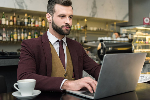 handsome businessman sitting at table with cup of coffee and typing on laptop in restaurant - Foto, imagen