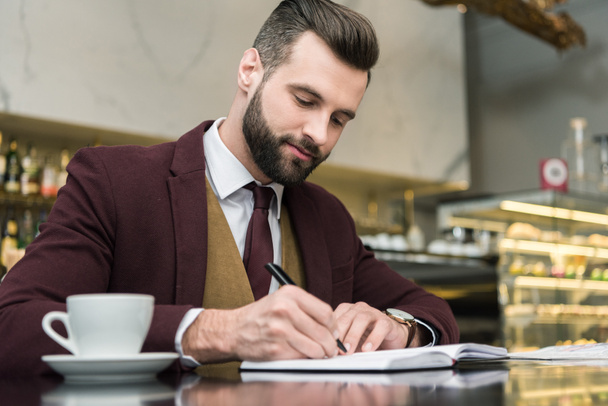 handsome businessman in formal wear sitting and writing in notebook at table in restaurant - Фото, изображение
