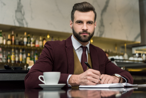 businessman in formal wear sitting, looking at camera and writing in notebook at table in restaurant - Foto, Imagem