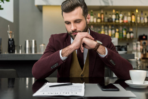 hombre de negocios pensativo en ropa formal con las manos dobladas sentadas en la mesa y mirando el contrato
 - Foto, imagen