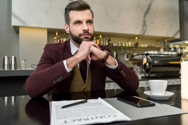 задумчивый бизнесмен сидит за столом с контрактом, кофе и смартфоном, глядя в сторону
 - Фото, изображение