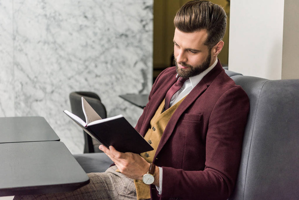 handsome businessman in formal wear sitting at restaurant and looking at notebook - Фото, изображение