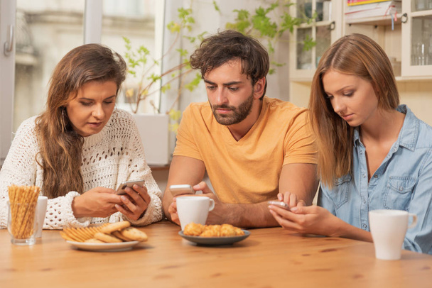 Friends sitting in the kitchen and watching on their phones. Internet browsing, and technology addiction concept - Foto, imagen