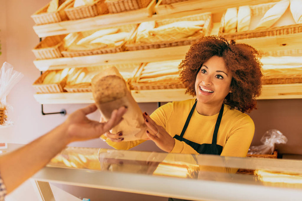 Młoda kobieta afro piekarz w piekarni, dając kija chleb zbożowe do klienta, uśmiechnięta i szczęśliwa. - Zdjęcie, obraz