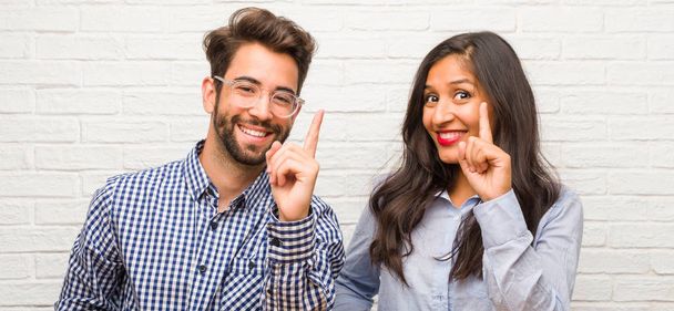 Young indian woman and caucasian man couple showing number one, symbol of counting, concept of mathematics, confident and cheerful - Photo, Image