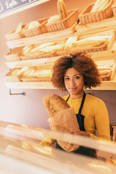 Młoda kobieta afro piekarz praca, trzymając kija chleb zbożowe, czuje ładne, bo ona kocha swoją pracę. - Zdjęcie, obraz