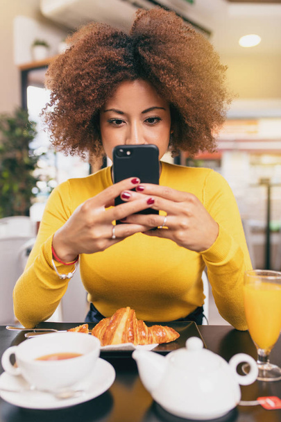 Νεαρή γυναίκα Αφρο γραπτά μηνύματα με το κινητό της τηλέφωνο στο καφέ, πίνοντας τσάι και τρώγοντας ένα κρουασάν. - Φωτογραφία, εικόνα