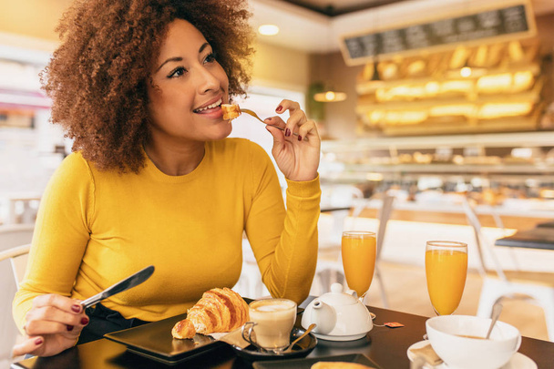 Młoda kobieta afro, śniadanie, jedzenia rogalika i picie kawy i soku pomarańczowego. - Zdjęcie, obraz