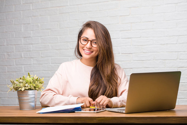 Porträt einer jungen Lateinstudentin, die fröhlich und mit einem breiten Lächeln auf ihrem Schreibtisch sitzt, selbstbewusst, freundlich und aufrichtig, die Positivität und Erfolg ausdrückt - Foto, Bild