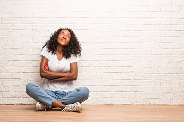 Jonge zwarte vrouw zitten op een houten vloer opzoeken, denk aan iets leuks en een idee, concept van verbeelding, blij en opgewonden - Foto, afbeelding