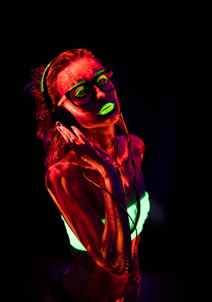 Портрет красивої молодої сексуальної дівчини в навушниках з ультрафіолетовою фарбою на її тілі. Красива жінка з сяючим бодіарт в чорному ламповому світлі
 - Фото, зображення