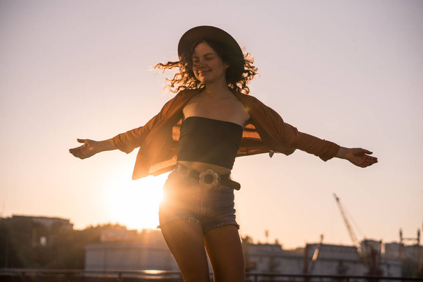 Curly chestnut-haired sexy girl in jeans shorts and hat is spinning on the sea port broadway at sunset, in industrial urban environment - 写真・画像
