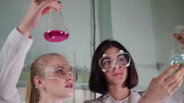 化学実験室。2 つの若い技術者は、自分たちの手でそれを見てテーブルからフラスコを取ります。肖像画 - 映像、動画