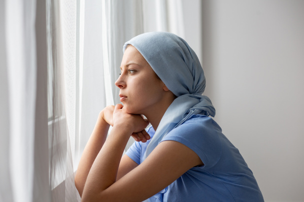 Thoughtful young girl suffering from ovarian cancer, wearing blue headscarf and looking through the window in medical center - Fotoğraf, Görsel
