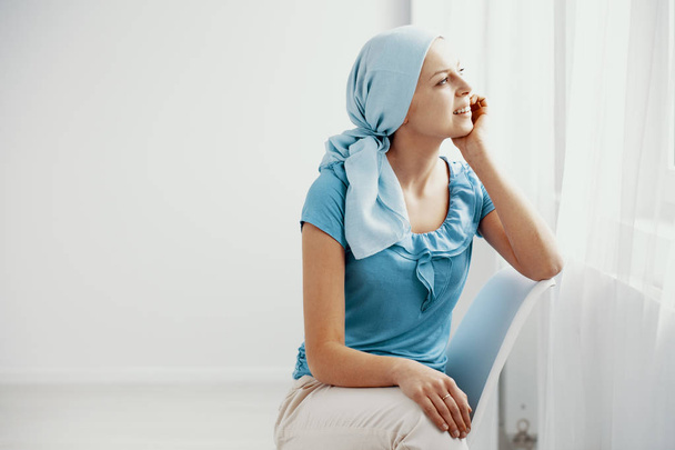 Smiling young girl suffering from lupus, wearing blue headscarf and looking through the window in hospital, copy space on the empty wall - Fotografie, Obrázek