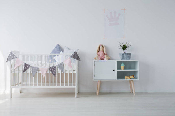 White wooden crib with pillows next to wooden cabinet with toy and green pant in grey, copy space on empty white wall - Photo, Image