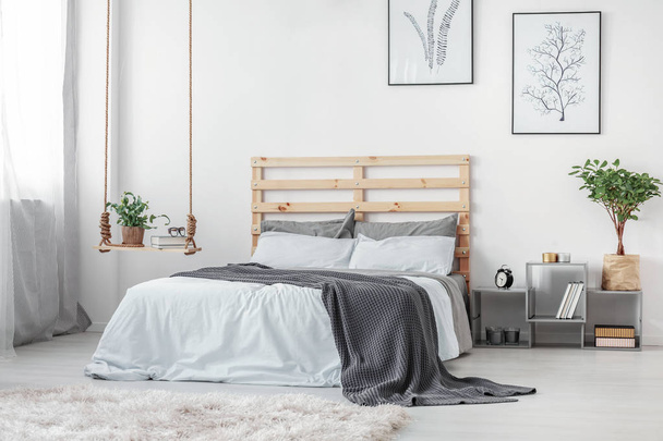 Stylish swing with books and plant in pot next to king size bed with wooden headboard and grey and white bedding - Photo, image