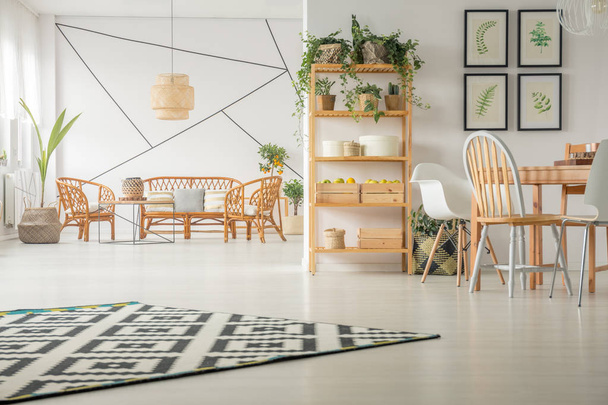 Apartamento de planta abierta interior con comedor y sala de estar con muebles de madera natural, galería de carteles florales en la pared y alfombra estampada en blanco y negro en el suelo
 - Foto, Imagen