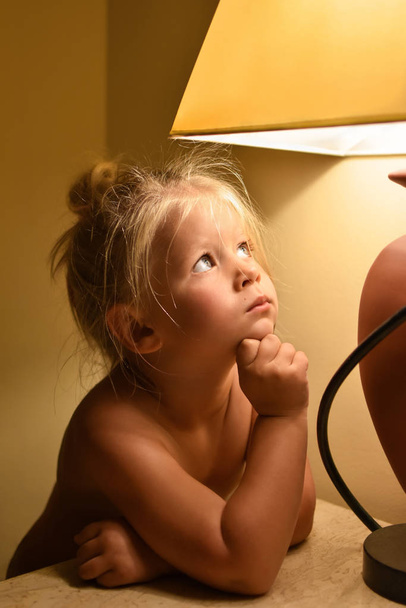 A child girl without clothes looks at the light from the lamp and dreams in the room in the evening. - Photo, Image