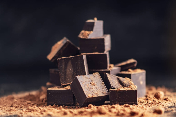 Pieces of dark bitter chocolate with cocoa powder on dark wooden background. The concept of confectionery ingredients - Fotoğraf, Görsel