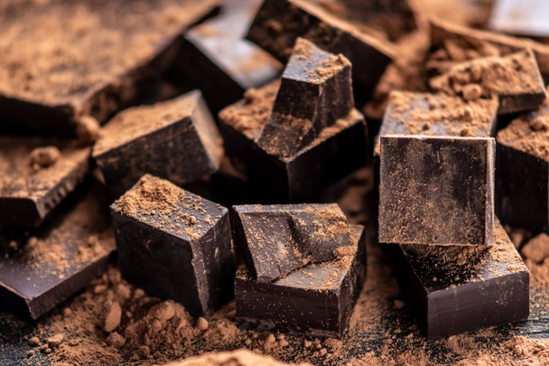 Pieces of dark bitter chocolate with cocoa powder on dark wooden background. The concept of confectionery ingredients - Fotografie, Obrázek