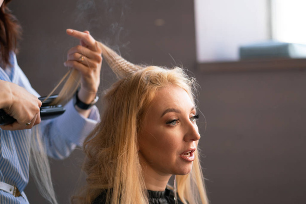 hermosa cliente rubia adulta se sienta en una silla, mientras que un maestro de peluquería en el salón hace su cabello con una ondulación y una permanente
 - Foto, Imagen