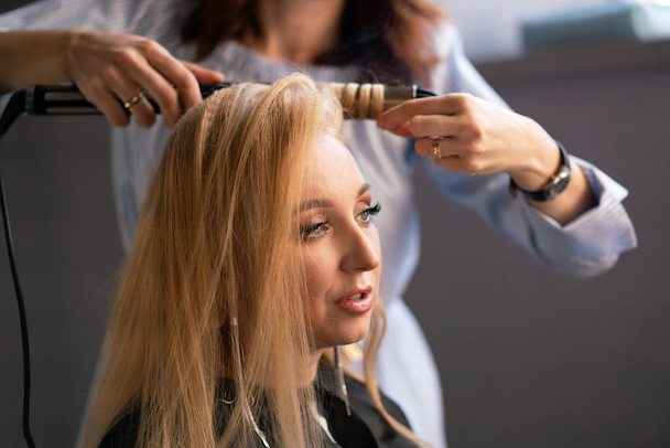 schöne erwachsene blonde Kundin sitzt auf einem Stuhl, während ein Friseurmeister im Salon ihr Haar mit Wellpappe und Dauerwelle frisiert - Foto, Bild