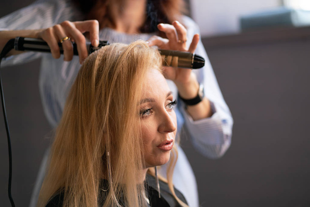 hermosa cliente rubia adulta se sienta en una silla, mientras que un maestro de peluquería en el salón hace su cabello con una ondulación y una permanente
 - Foto, imagen