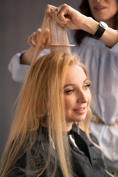prachtige volwassen blonde client zit in een stoel, terwijl een kapper-meester in de salon haar haren met een vouw- en een permanent doet - Foto, afbeelding