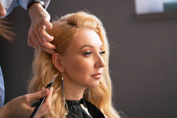 prachtige volwassen blonde client zit in een stoel, terwijl een kapper-meester in de salon haar haren met een vouw- en een permanent doet - Foto, afbeelding