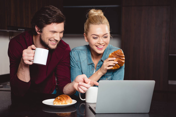 hombre guapo señalando con el dedo en la pantalla del ordenador portátil mientras chica atractiva sosteniendo croissant en la cocina
 - Foto, imagen