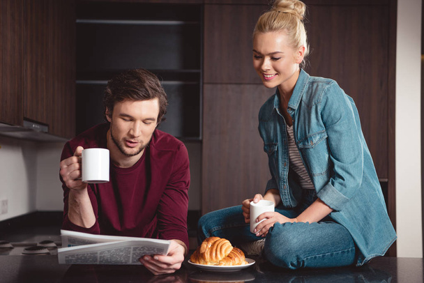 pohledný muž držící šálek kávy a čtenářský deník zároveň atraktivní dívka seděla na stole a usmívá se v kuchyni - Fotografie, Obrázek
