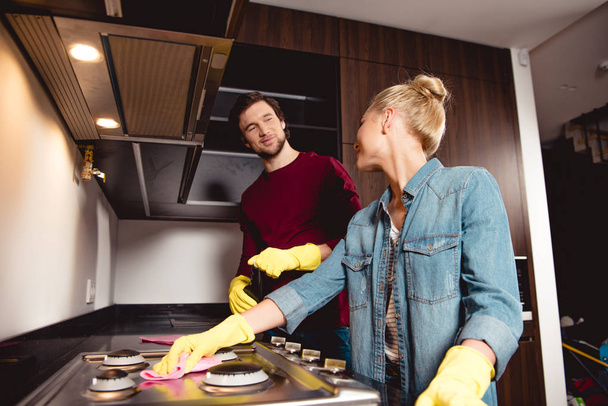 smiling couple in rubber gloves cleaning kitchen and furniture together and looking at each other - Foto, Bild