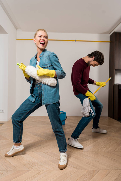 cheerful family couple in rubber gloves husband performing with mop and duster in apartment - Photo, Image