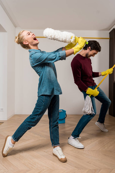 happy girl in rubber gloves singing with duster while husband performing with mop in apartment - Foto, imagen